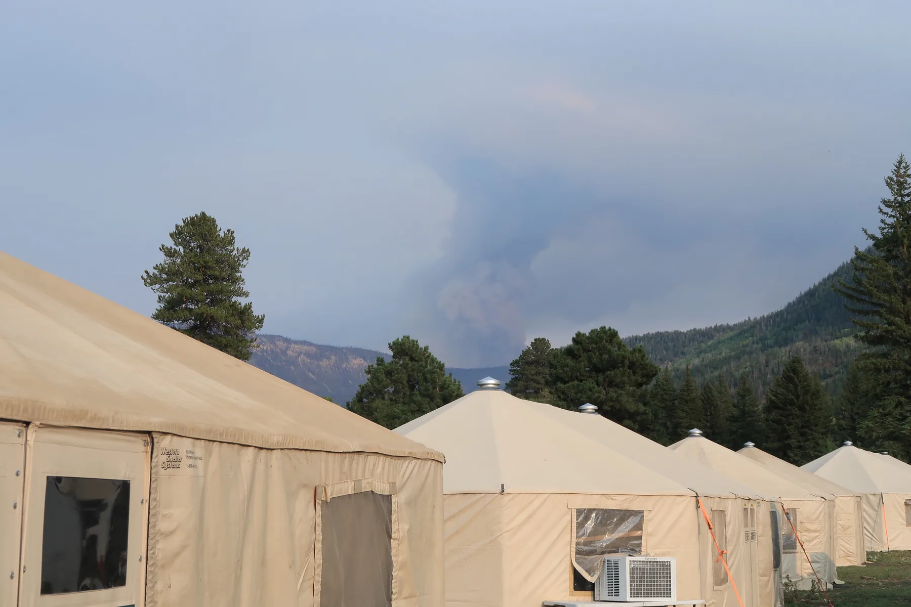 A row of Evergreen Resource yurts with a wildfire smoke plume on the mountain behind the camp
